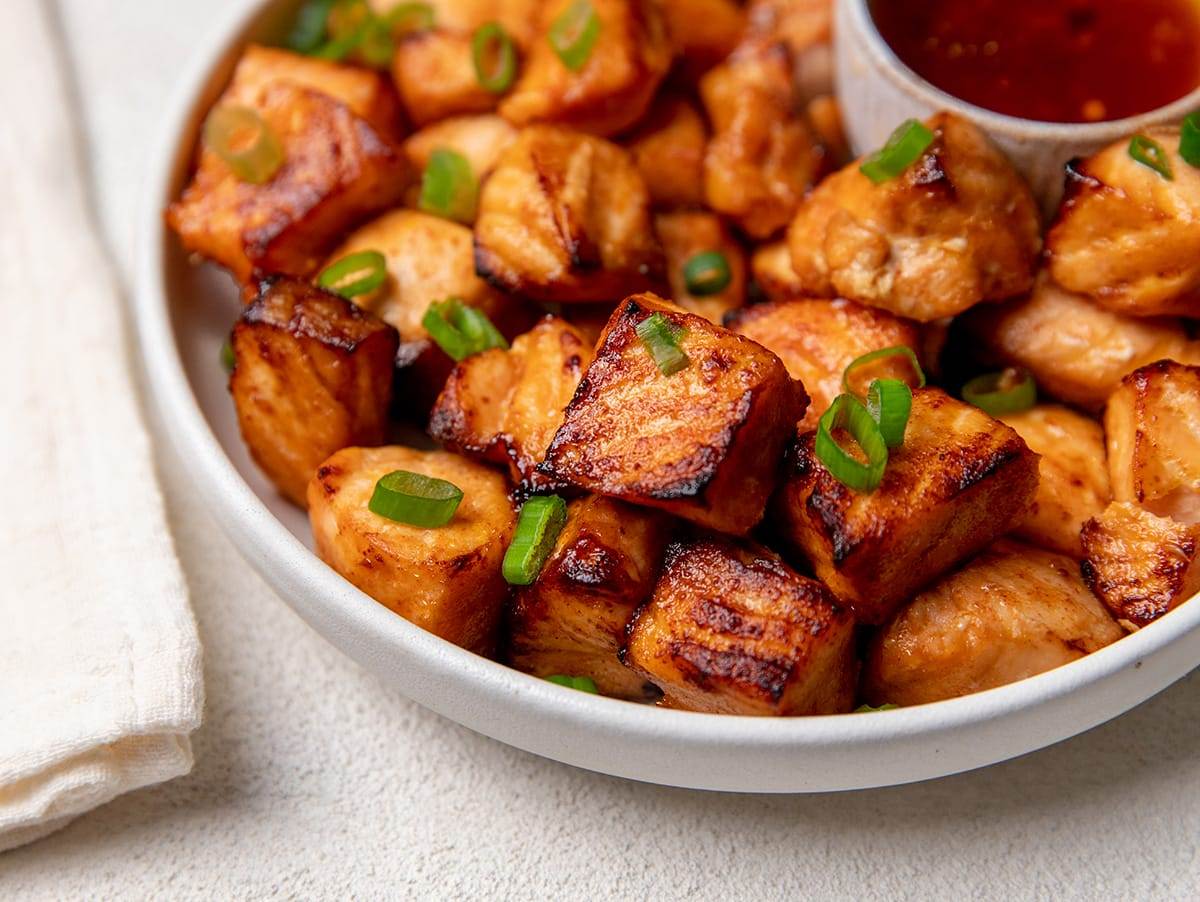 A bowl of crispy air fryer salmon bites coated in glossy garlic butter sauce with chopped parsley, photographed on a kitchen counter