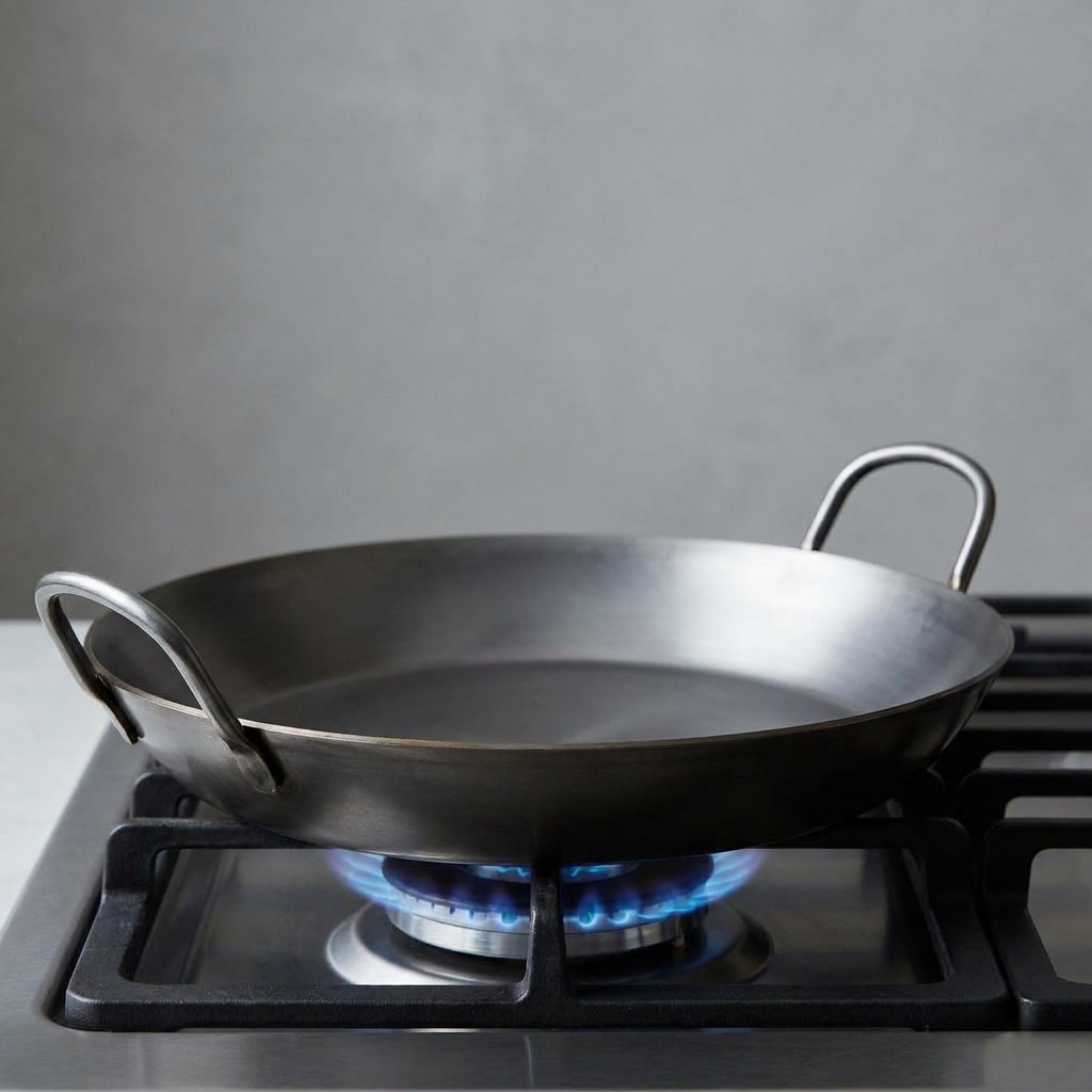 A clean carbon steel paella pan centered on a stovetop burner, showing its wide shallow shape and two loop handles