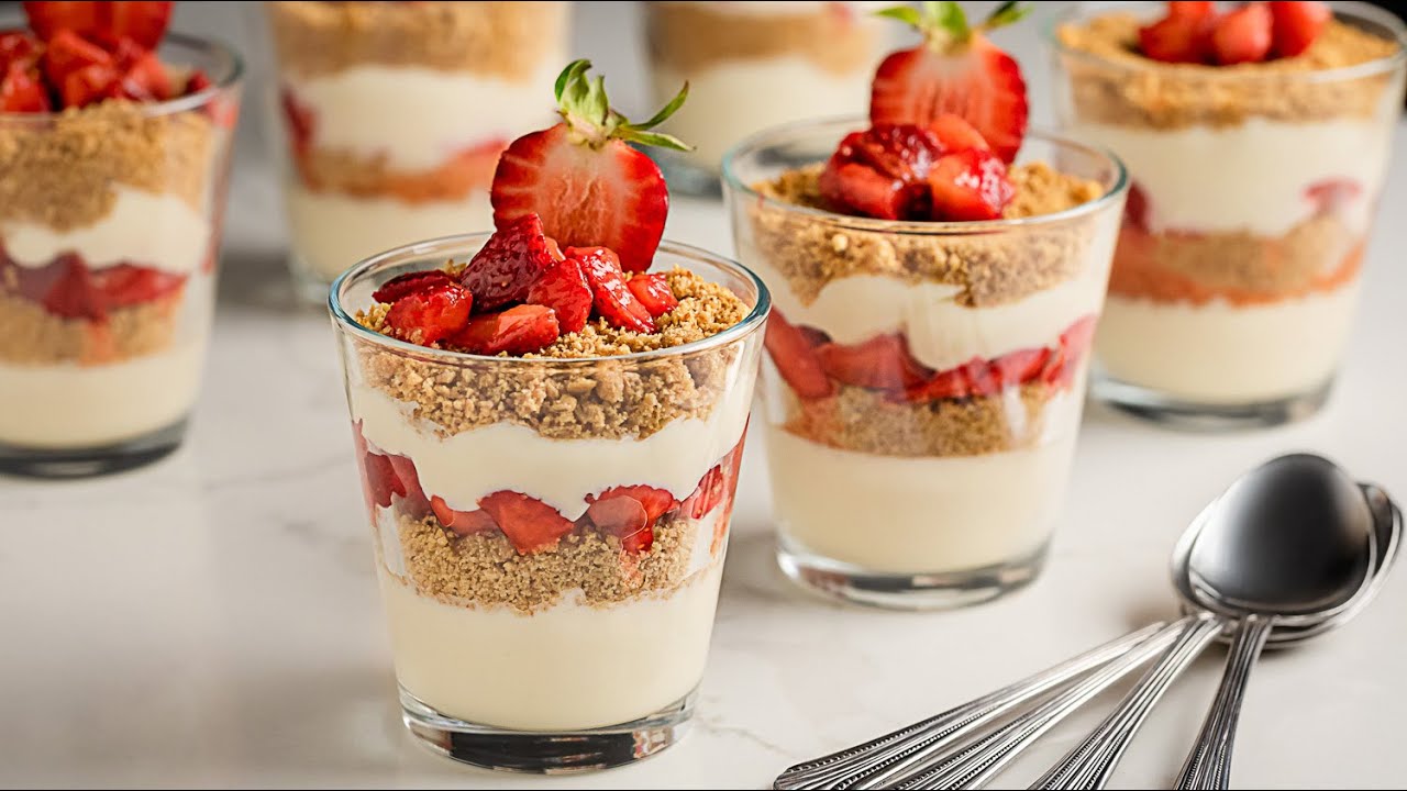 A clear glass cup filled with layers of strawberry cheesecake yogurt and graham cracker crumbs, topped with sliced strawberries