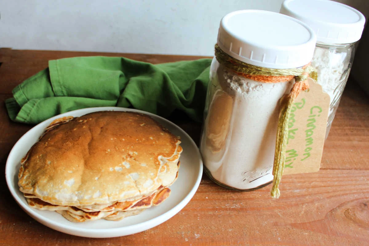 A glass mason jar filled with homemade pancake mix on a bright kitchen counter, with a wooden spoon and a small bowl of flour nearby