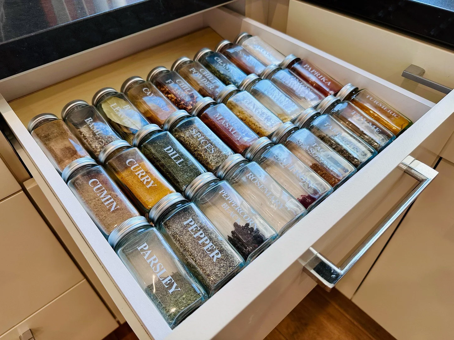 A kitchen spice drawer with small labeled glass jars, including one jar of reddish taco seasoning tucked beside cumin, paprika, and chili powder