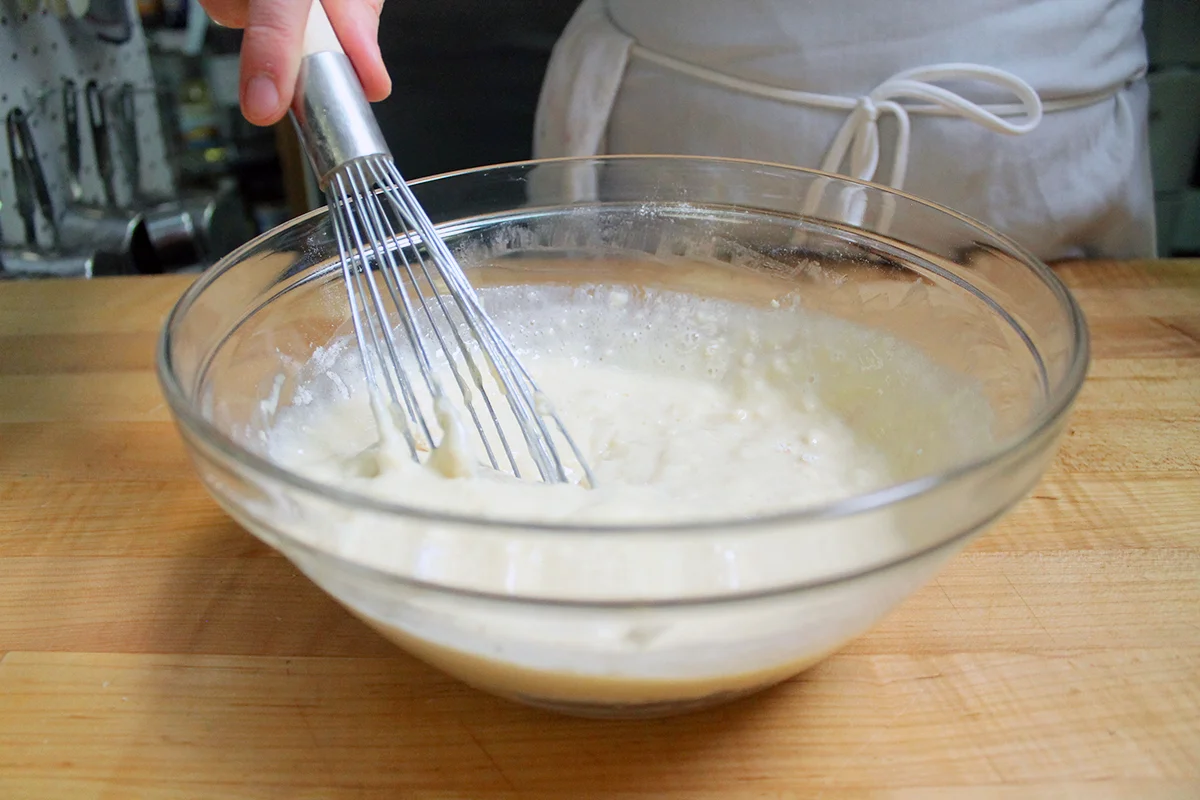 A large mixing bowl with flour-based pancake mix being whisked thoroughly, with measuring spoons resting on the counter