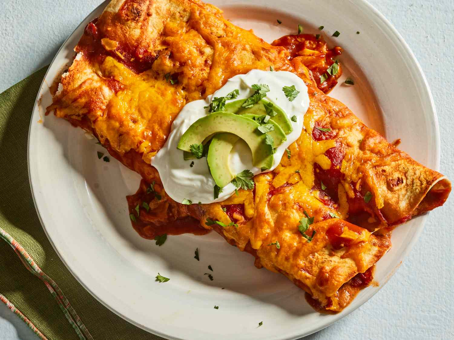 A plated serving of chicken enchiladas topped with sour cream and chopped cilantro with lime wedges on the side