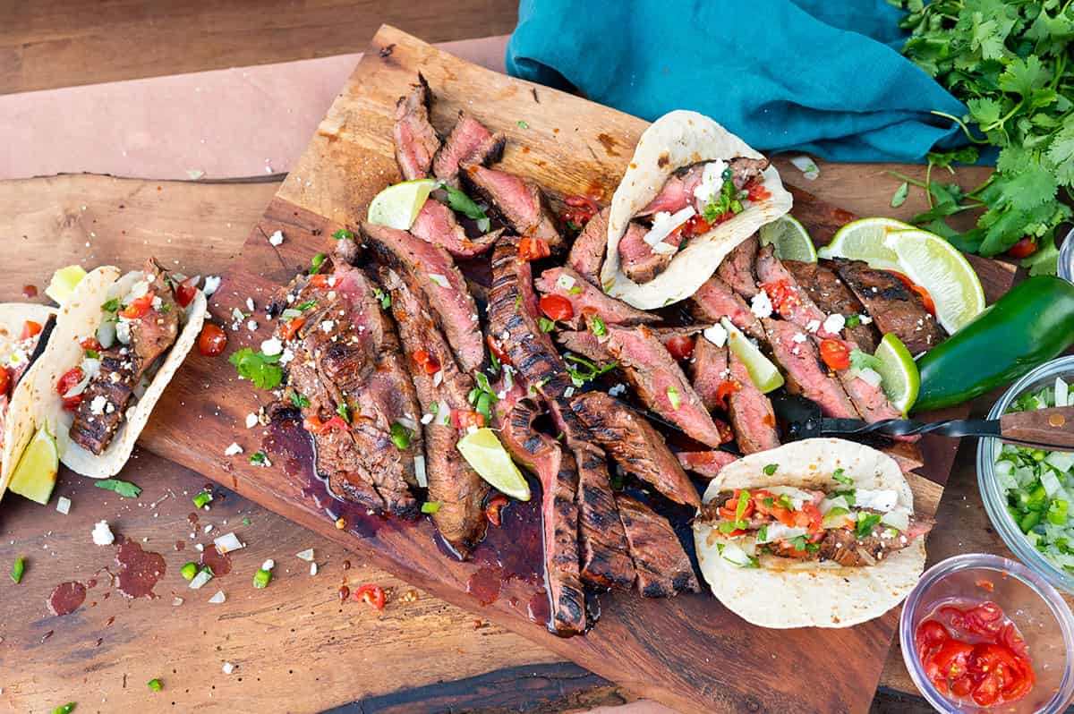 A platter of beef tacos in warm corn tortillas topped with lettuce, diced tomatoes, shredded cheese, and cilantro, with lime wedges on the side on a kitchen table