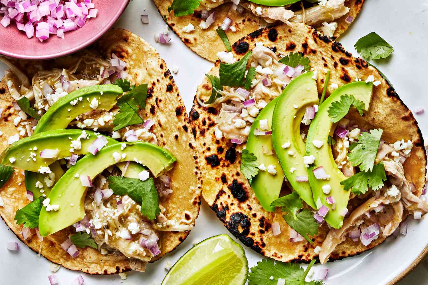 A platter of shredded salsa verde chicken with warm corn tortillas, diced onion, cilantro, and lime wedges on a family dinner table