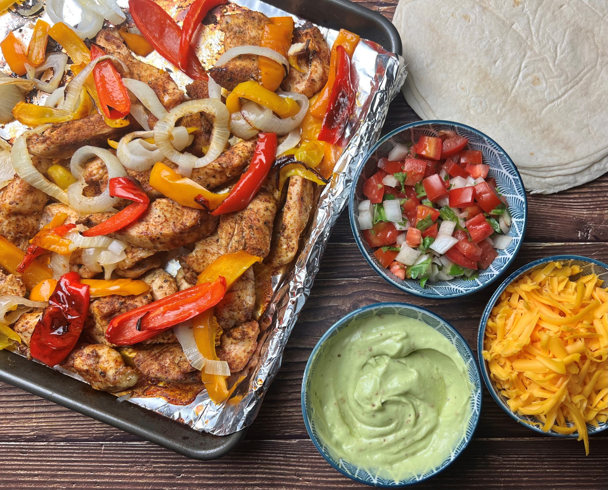 A rimmed sheet pan filled with sliced chicken, bell peppers, and onions roasted until lightly charred, with visible spices and a few lime wedges tucked into the pan