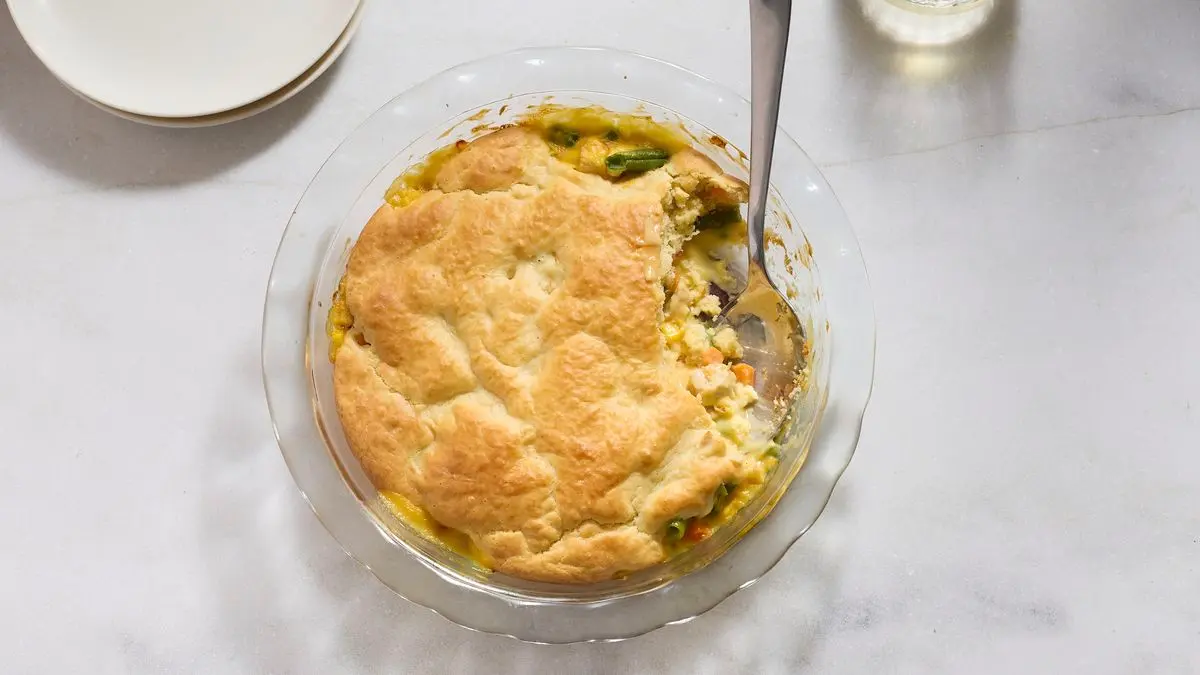 A serving of chicken pot pie on a plate with flaky puff pastry on top and creamy chicken, peas, and carrots underneath, photographed at a dinner table