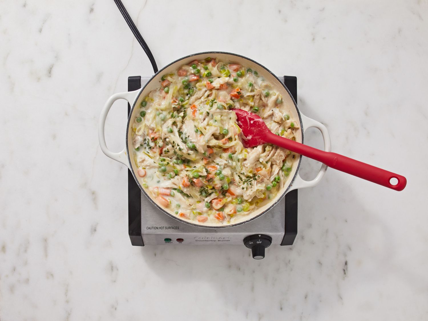 A skillet filled with creamy chicken pot pie filling with visible carrots, celery, peas, and shredded chicken being stirred with a wooden spoon