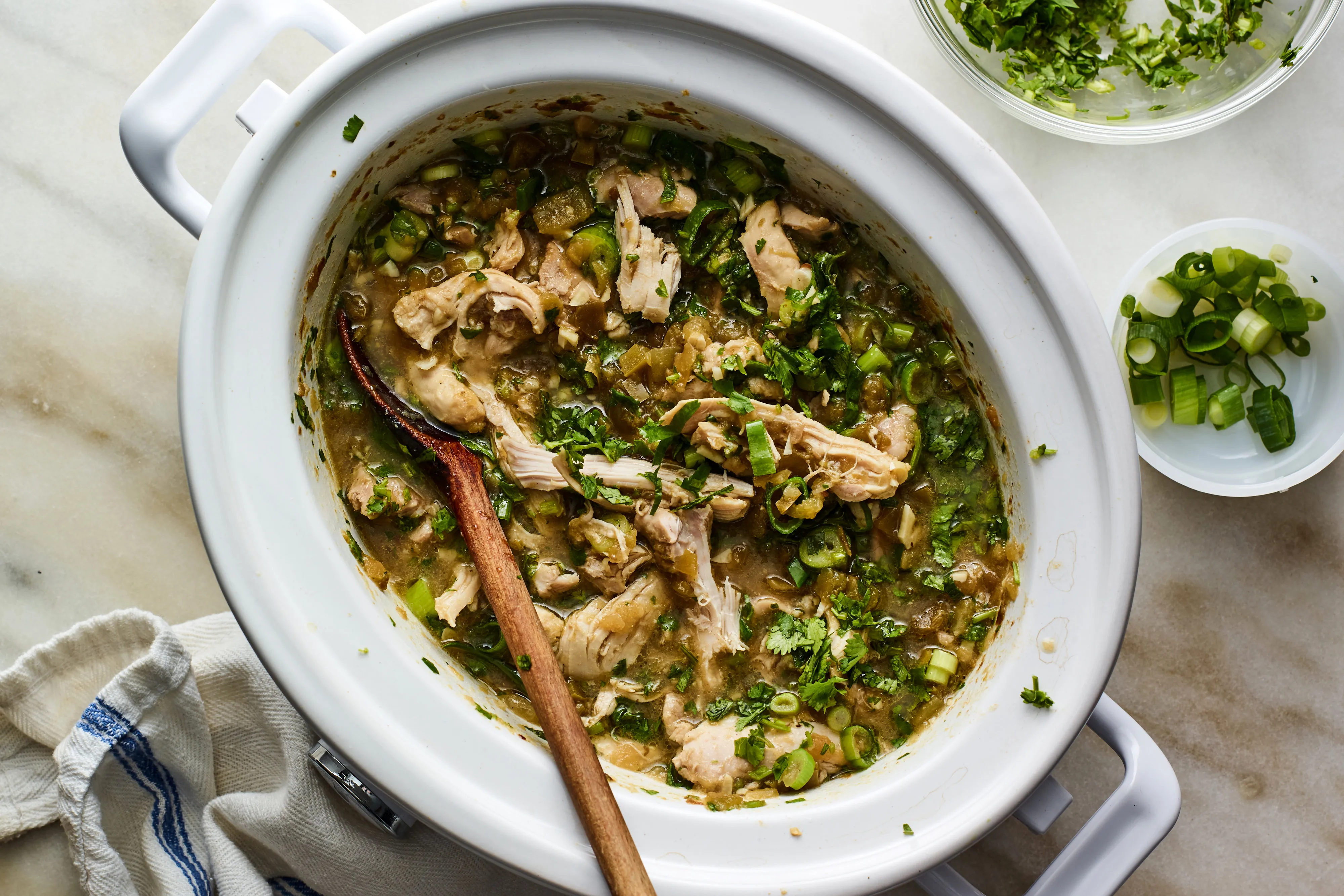 A slow cooker insert filled with shredded salsa verde chicken being stirred with a wooden spoon, with green sauce pooling around the meat