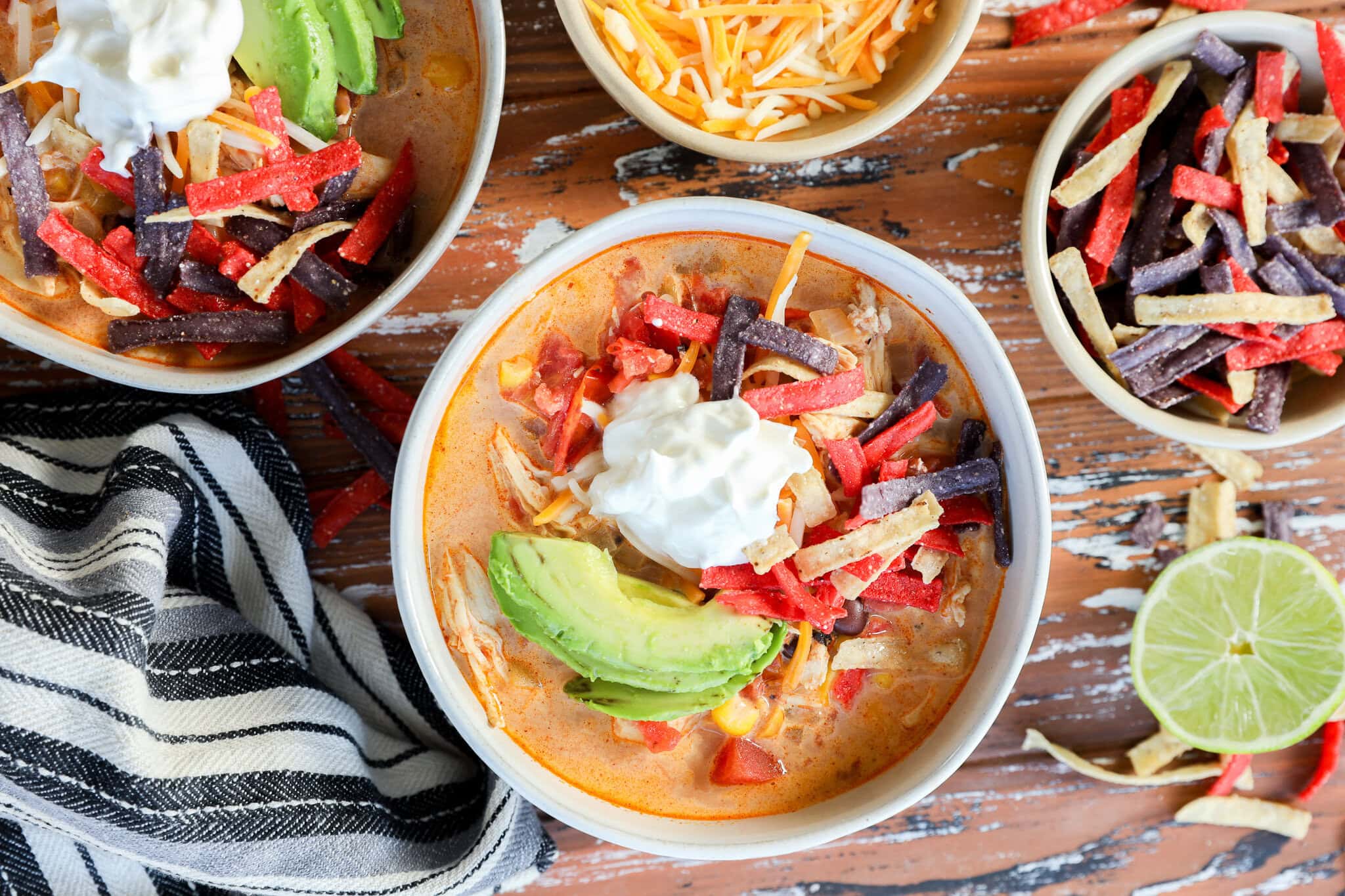 A steaming bowl of creamy chicken tortilla soup topped with crispy tortilla strips, diced avocado, cilantro, and a lime wedge on a wooden table