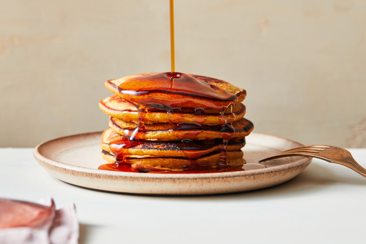 A tall stack of fluffy pancakes on a plate with maple syrup being poured over the top, with a fork resting beside the plate