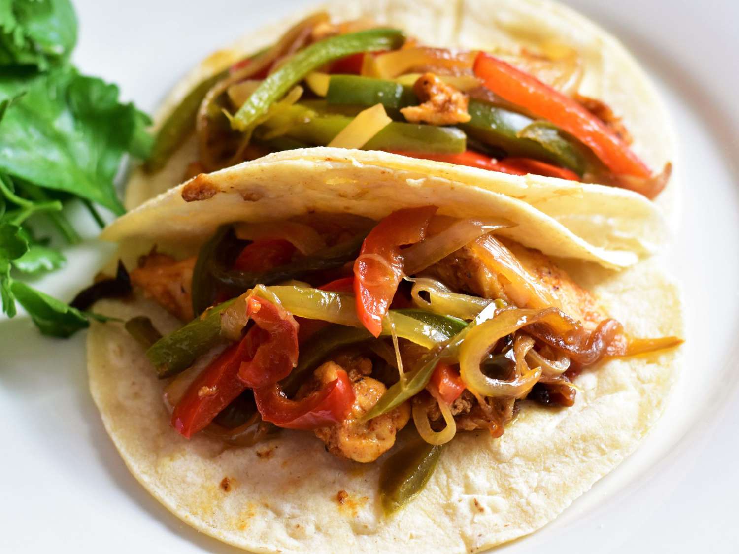 Chicken and roasted peppers and onions being spooned from a sheet pan into warm flour tortillas on a wooden cutting board