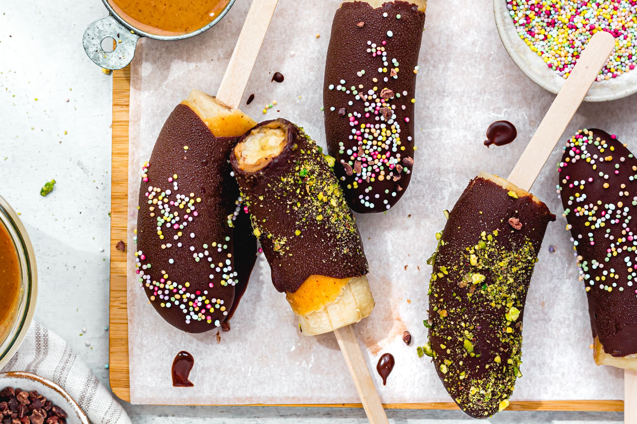 Chocolate-dipped banana pops resting on parchment paper with a few chopped nuts scattered nearby