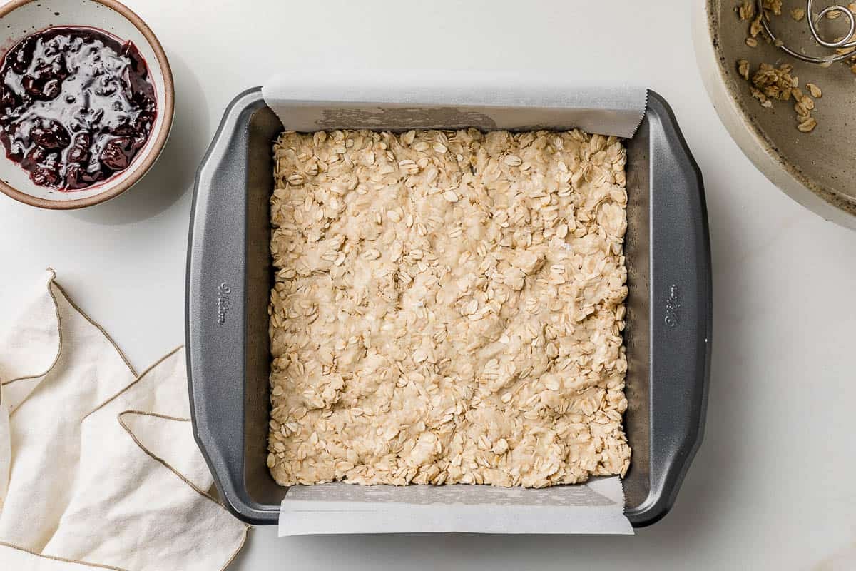 Hands pressing a peanut butter oat mixture firmly into a parchment-lined square baking pan