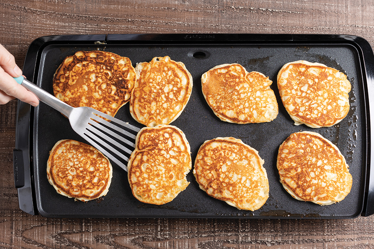 Several pancakes cooking on a warm griddle with bubbles forming on the surface and a spatula nearby
